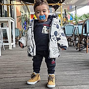 Imran a rejoint le concours — aidez-le/la à gagner de superbes lots ! toddler, child, pacifier, jacket, mickey_mouse, outdoor, deck, wooden_floor, string_lights, cafe, chairs, tables, boots, clothing, smile, person, standing, casual, daylight, cute