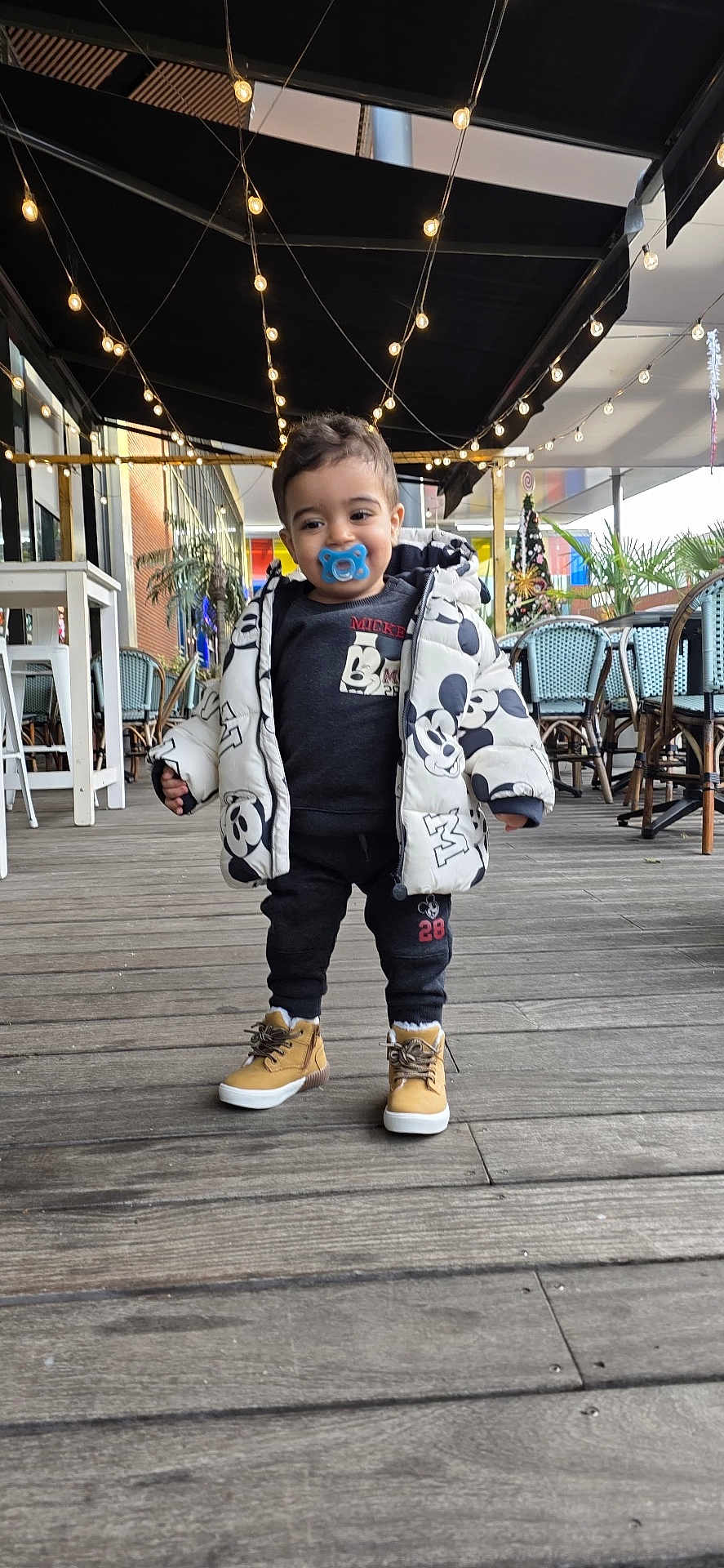 Imran a rejoint le concours — aidez-le/la à gagner de superbes lots ! toddler, child, pacifier, jacket, mickey_mouse, outdoor, deck, wooden_floor, string_lights, cafe, chairs, tables, boots, clothing, smile, person, standing, casual, daylight, cute