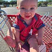 Hudson is registered to the contest to win money with this photo: baby, child, shopping_cart, red_shirt, smiling, outdoor, parking_lot, sunlight, happy, face, foot, hand, toy_clip, phone_case, metal, retail, summer, casual_clothing, infant, playful