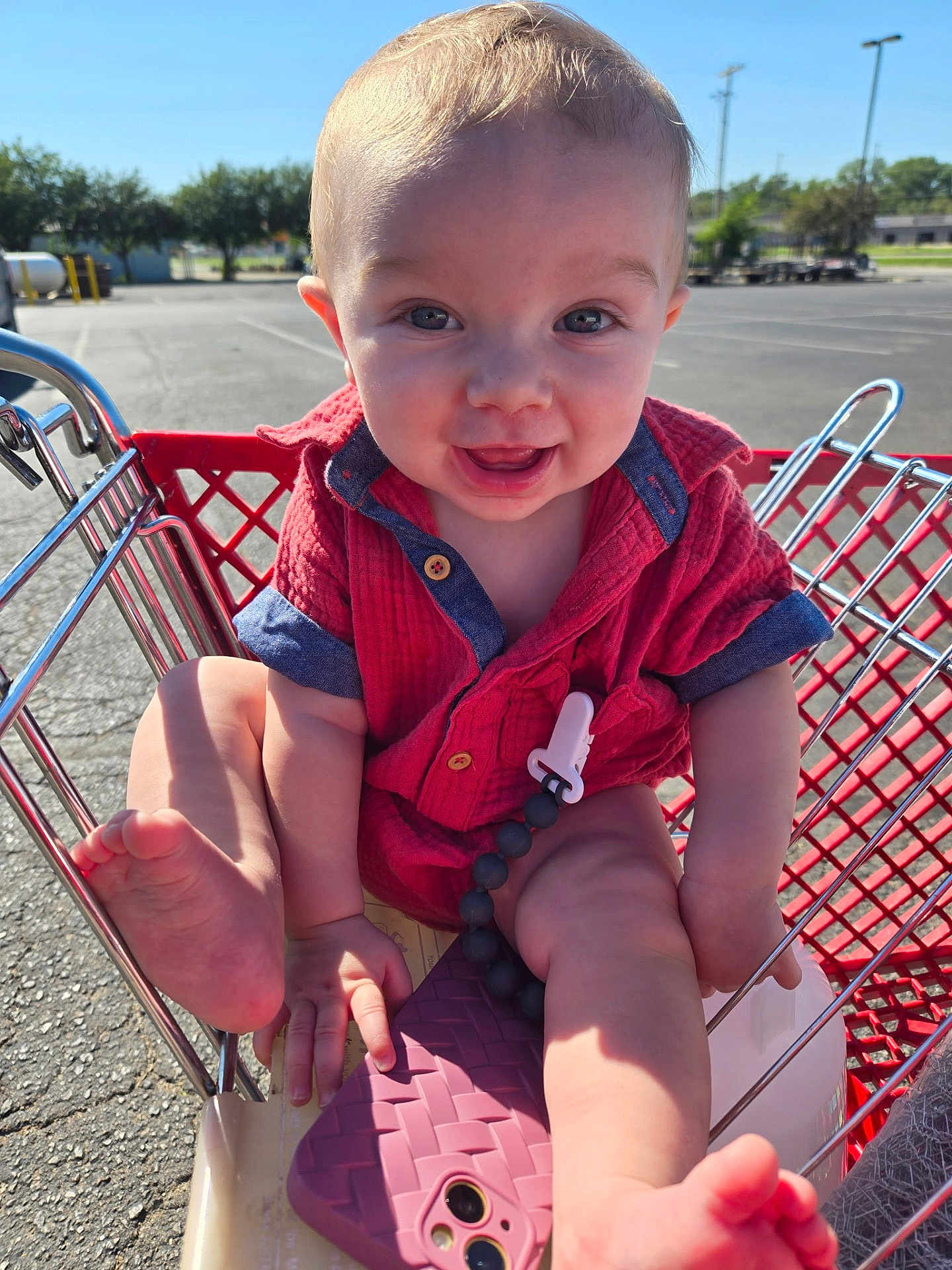 Hudson is registered to the contest to win money with this photo: baby, child, shopping_cart, red_shirt, smiling, outdoor, parking_lot, sunlight, happy, face, foot, hand, toy_clip, phone_case, metal, retail, summer, casual_clothing, infant, playful