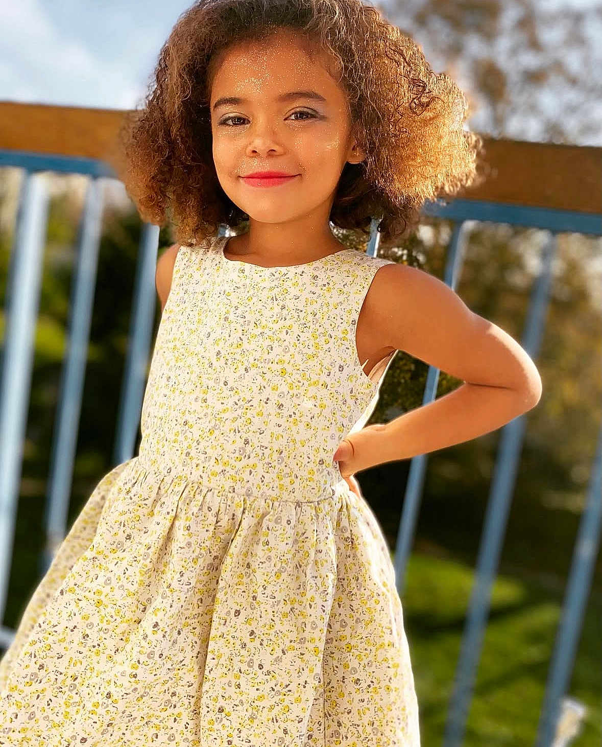 Maya participe au concours pour gagner de l'argent avec cette photo : child, girl, curly_hair, smiling, dress, floral_pattern, outdoor, sunlight, fence, greenery, portrait, standing, happy, confident, fashion, summer, nature, person, daylight, casual