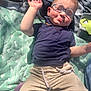 toddler, child, glasses, blue_shirt, beige_pants, blanket, green_blanket, outdoor, sunlight, hand_up, face, closed_eyes, cute, playing, relaxing, person, baby, casual_clothing, summer, smiling