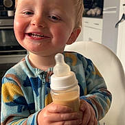 Reagan is registered to the contest to win money with this photo: toddler, child, baby_bottle, smiling, happy, high_chair, kitchen, blond_hair, rosy_cheeks, casual_clothing, indoor, person, holding, drink, cute, young_child, furniture, portrait, feeding, playful
