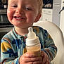 toddler, child, baby_bottle, smiling, happy, high_chair, kitchen, blond_hair, rosy_cheeks, casual_clothing, indoor, person, holding, drink, cute, young_child, furniture, portrait, feeding, playful