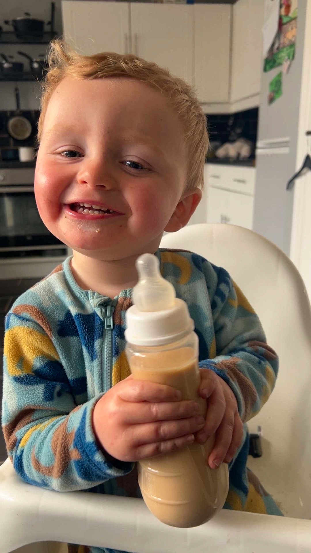 Reagan is registered to the contest to win money with this photo: toddler, child, baby_bottle, smiling, happy, high_chair, kitchen, blond_hair, rosy_cheeks, casual_clothing, indoor, person, holding, drink, cute, young_child, furniture, portrait, feeding, playful
