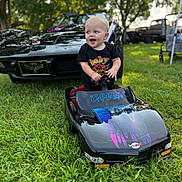 Cam is registered to the contest to win money with this photo: toddler, child, toy_car, miniature_car, black_car, vintage_car, grass, outdoor, daylight, trees, smiling, happy, steering_wheel, vehicle, parked_car, person, nature, greenery, play, casual_clothing