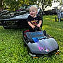 toddler, child, toy_car, miniature_car, black_car, vintage_car, grass, outdoor, daylight, trees, smiling, happy, steering_wheel, vehicle, parked_car, person, nature, greenery, play, casual_clothing