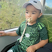 Idris participe au concours pour gagner de l'argent avec cette photo : child, pacifier, cap, green_shirt, shorts, sitting, window, indoor, bag, calm, face, young_boy, portrait, patterned_clothing, daylight, seat, hand, looking_away, casual, sportswear