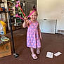 Brynlee Mae is registered to the contest to win money with this photo: boxes, casual_clothing, child, dress, floor, flower, girl, hand, happy, indoor, person, pink_crocs, playful, purple_dress, room, shelves, smiling, standing, toy, young_child