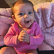 Nova is registered to the contest to win money with this photo: baby, infant, pink_onesie, blanket, clapping, smiling, happy, indoors, living_room, carpet, furniture, table, cozy, child, portrait, cute, hands, face, person, seated