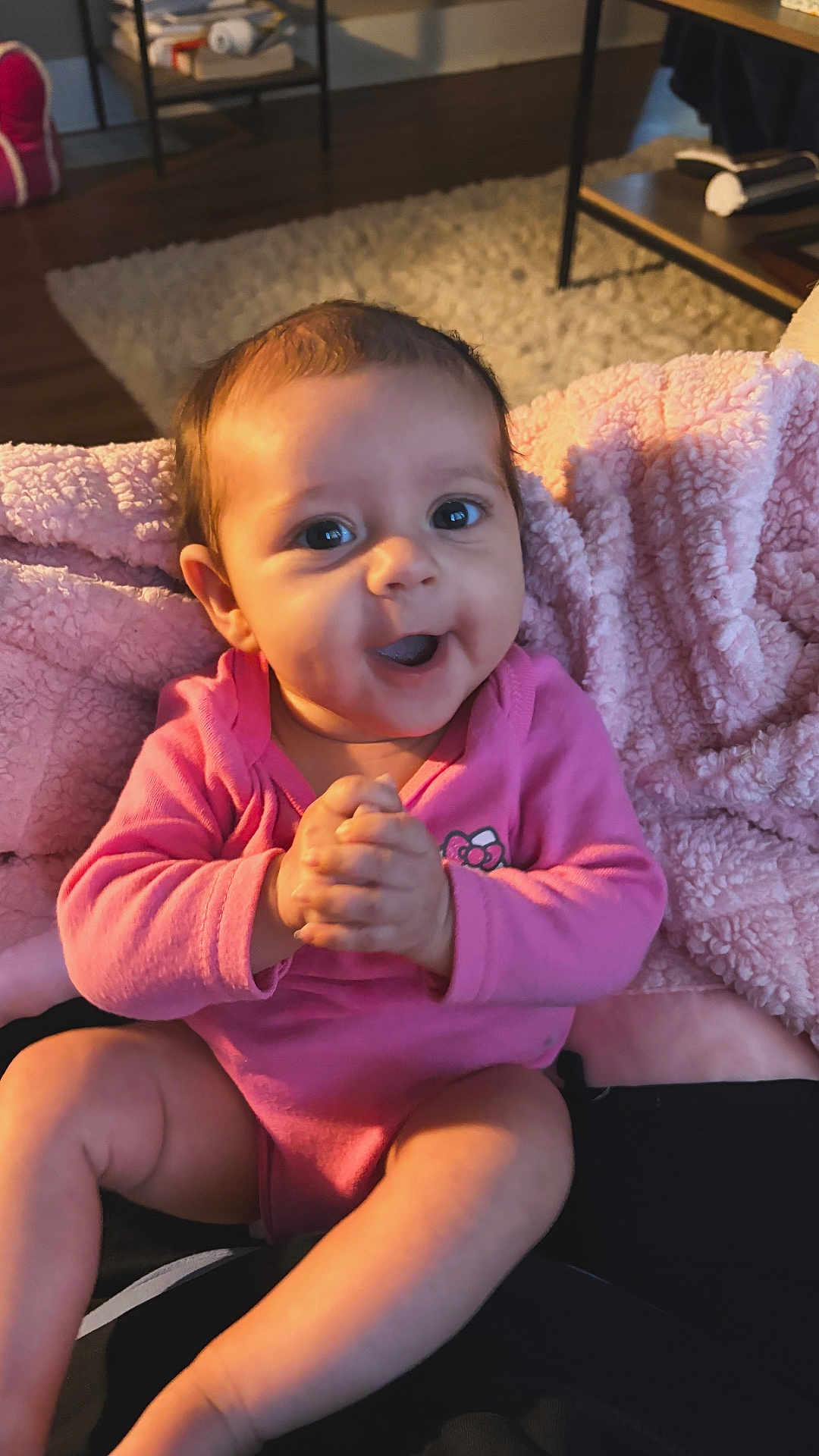 Nova is registered to the contest to win money with this photo: baby, infant, pink_onesie, blanket, clapping, smiling, happy, indoors, living_room, carpet, furniture, table, cozy, child, portrait, cute, hands, face, person, seated