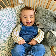 Andréas a rejoint le concours — aidez-le/la à gagner de superbes lots ! baby, child, smiling, denim_overalls, striped_shirt, sock, teddy_bear, stuffed_animal, wicker_chair, cushion, indoor, happy, cute, infant, person, portrait, seated, cozy, comfortable, playful
