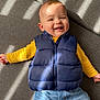 baby, blue_jeans, casual_clothing, child, couch, cute, face, happy, home, indoor, infant, lying_down, person, portrait, puffy_vest, relaxed, shadow, smiling, sunlight, yellow_shirt