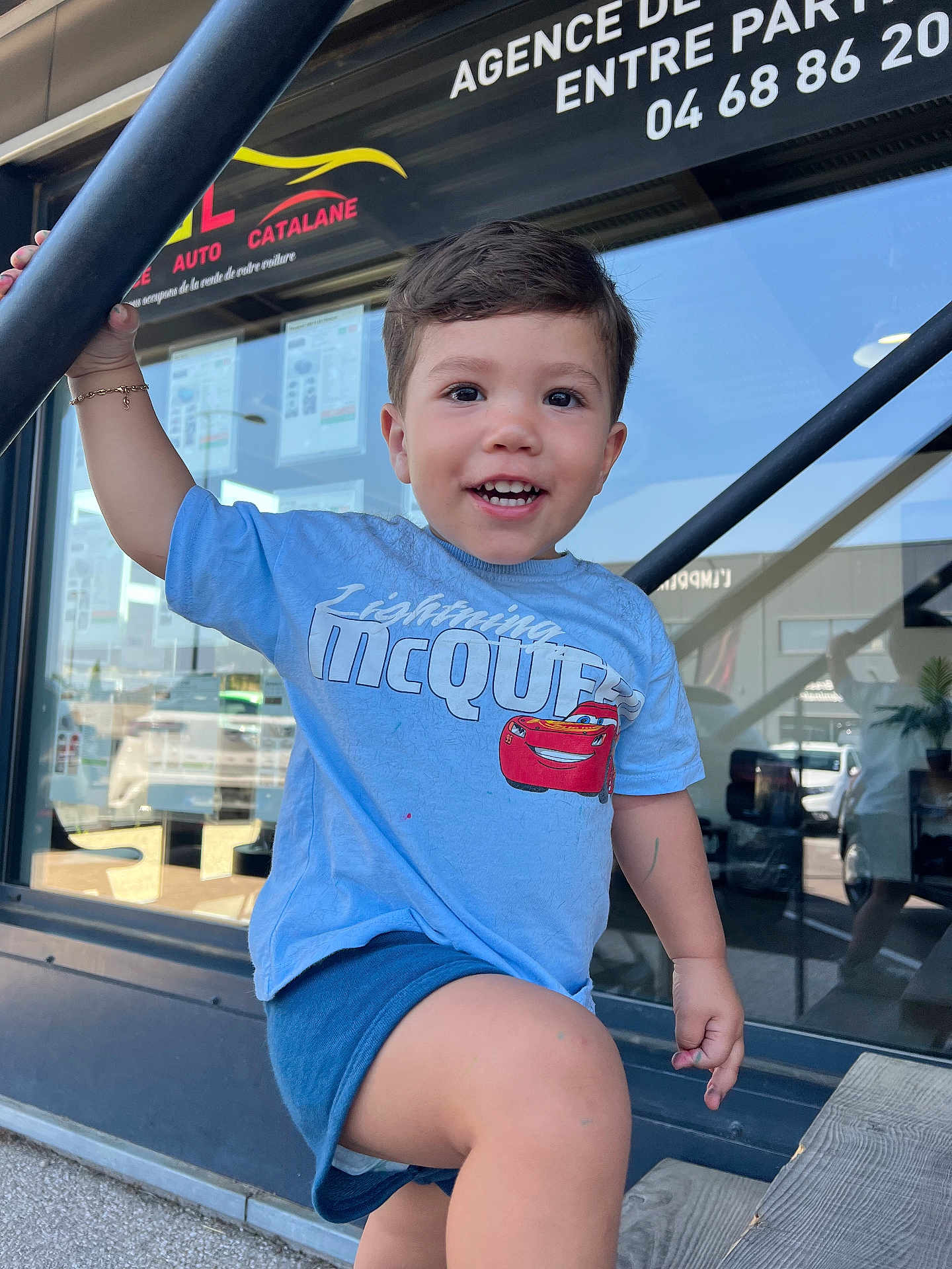 Battistu a rejoint le concours — aidez-le/la à gagner de superbes lots ! child, toddler, smiling, blue_shirt, shorts, outdoor, storefront, glass_window, reflection, metal_pole, daylight, happy, casual_clothing, boy, urban, sidewalk, hand, face, walking, sunlight
