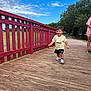 toddler, child, running, bridge, wooden_floor, red_railing, outdoor, park, trees, grass, blue_sky, clouds, adidas, shorts, tshirt, sneakers, person, happy, smiling, casual_clothing