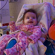 Jaylee is registered to the contest to win money with this photo: baby, child, infant, smiling, hospital, medical_equipment, bandage, pajamas, pink_clothing, blanket, chair, portrait, indoor, sitting, lying_down, raised_fist, balloon, colorful, happy, face