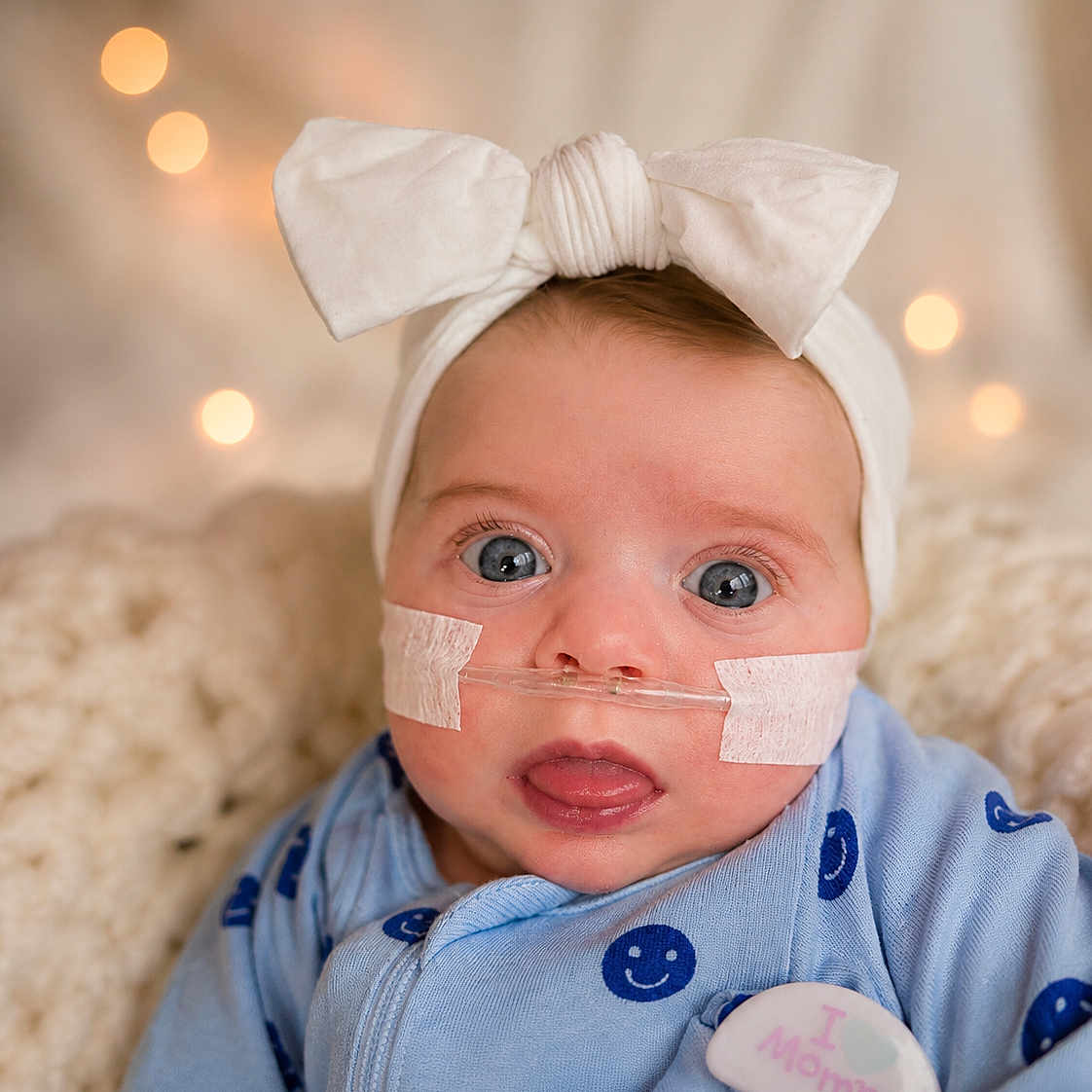 Jaylee joined the competition — help win amazing prizes! baby, infant, bow_headband, blue_outfit, smiley_face, nasal_oxygen_tube, tape, closeup, portrait, cute, wide_eyes, soft_background, bokeh, blanket, indoors, child, newborn, expression, wearing, headwear