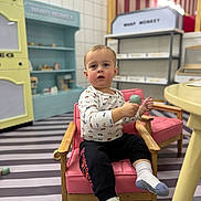 Rhett-Michael joined the competition — help win amazing prizes! toddler, child, playroom, indoor, pink_chair, wooden_armchair, toy, holding_toy, curious_expression, portrait, sitting, socks, striped_floor, play_kitchen, shelves, pastel_colors, bokeh_background, casual_clothing, retail_display, young_child
