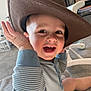 toddler, child, smiling, hat, cowboy_hat, onesie, striped_clothing, laughing, teeth, hand, indoor, carpet, blanket, baby_gear, closeup, portrait, playful, happy, seat, home