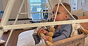 Rhett-Michael is registered to the contest to win money with this photo: baby, blanket, blinds, child, curtain, door, hanging_jumper, harness, infant, living_room, ottoman, play_gym, rug, socks, sofa, swing, toy, window, wood_floor, wooden_beads