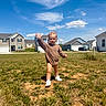toddler, child, grass, field, suburban, houses, blue_sky, clouds, smiling, waving, outdoor, sunny, casual_clothing, young_child, walking, daylight, residential_area, happy, bare_legs, shoes