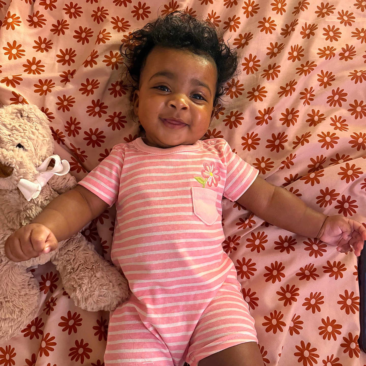 Kameryn is registered to the contest to win money with this photo: baby, baby_girl, blanket, child, curly_hair, cute, floral_pattern, happy, indoors, infant, lying_down, pajamas, pink_clothing, playful, portrait, smiling, soft_toys, striped_outfit, stuffed_animal, teddy_bear