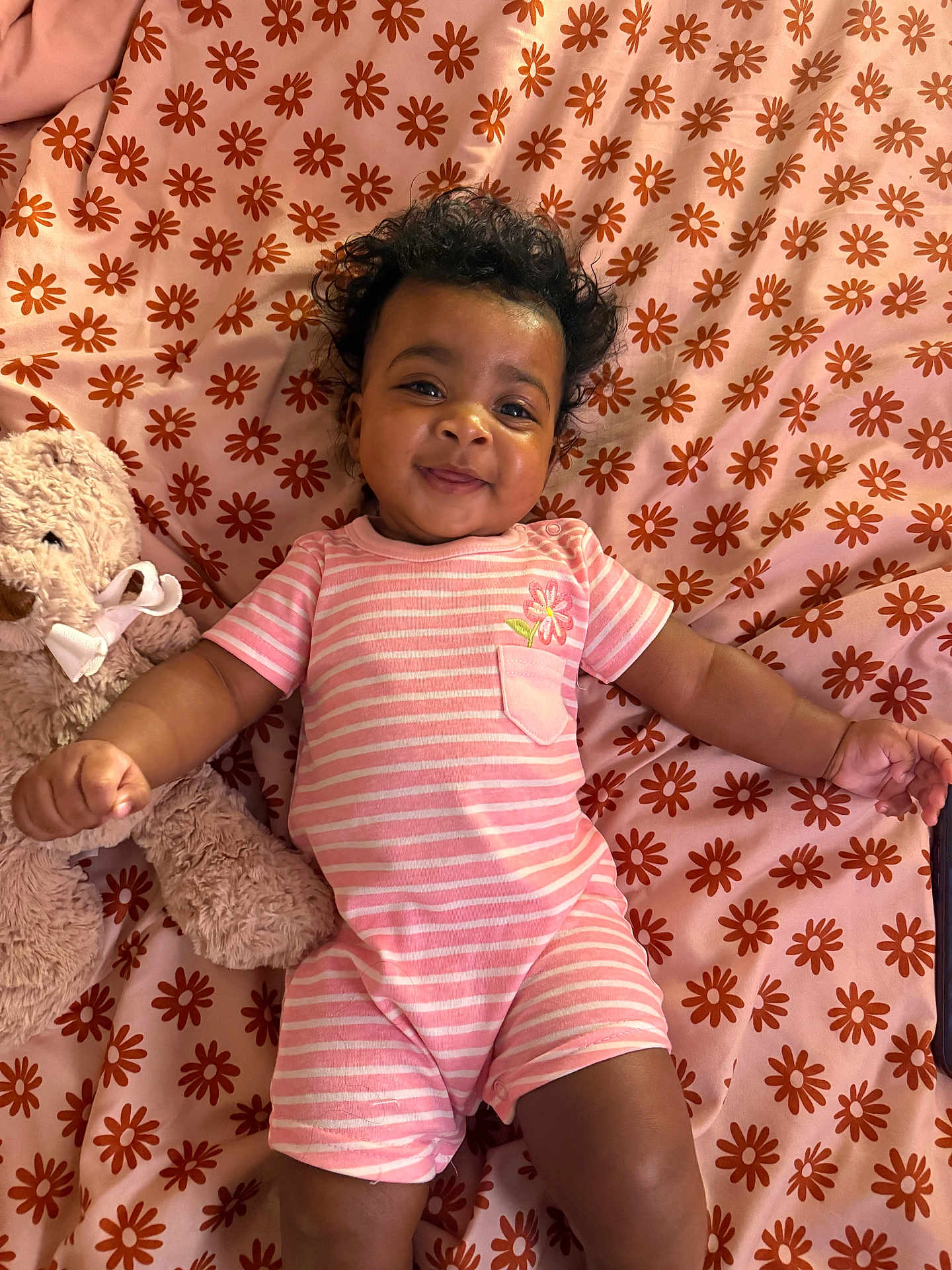Kameryn is registered to the contest to win money with this photo: baby, infant, smiling, curly_hair, pink_clothing, striped_outfit, teddy_bear, stuffed_animal, blanket, floral_pattern, lying_down, cute, child, indoors, happy, portrait, soft_toys, baby_girl, pajamas, playful