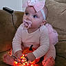 baby, child, pacifier, headband, bow, pink_clothing, pumpkin, string_lights, glowing_lights, couch, indoors, cute, blue_eyes, toddler, sitting, barefoot, decor, holiday, soft_lighting, curious