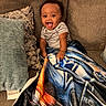 Idris is registered to the contest to win money with this photo: baby, child, infant, sitting, couch, blanket, pillow, striped_shirt, smiling, tongue_out, happy, portrait, indoors, home, furniture, textile, cozy, playful, photograph, facial_expression