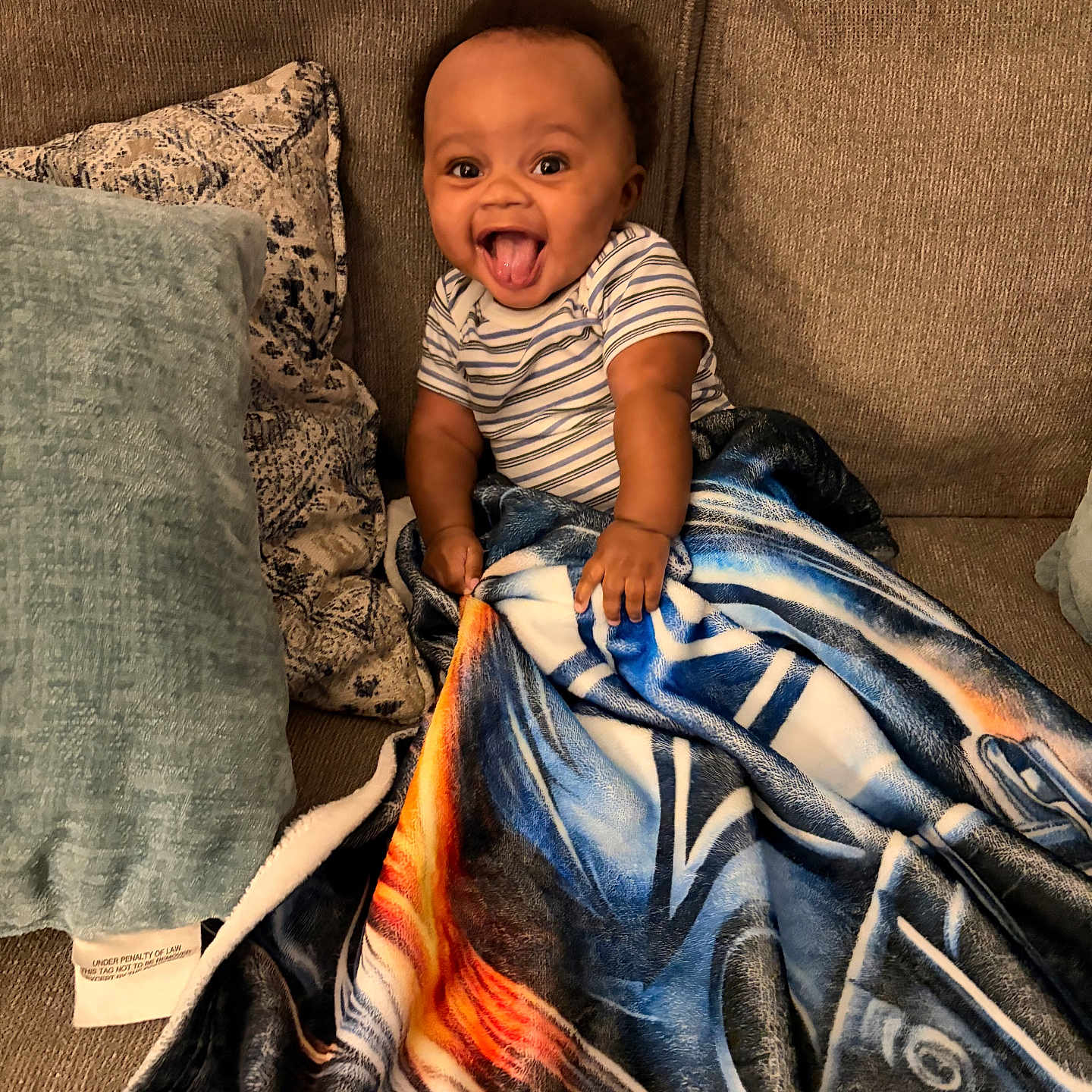 Idris is registered to the contest to win money with this photo: baby, child, infant, sitting, couch, blanket, pillow, striped_shirt, smiling, tongue_out, happy, portrait, indoors, home, furniture, textile, cozy, playful, photograph, facial_expression