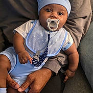 Idris joined the competition — help win amazing prizes! baby, infant, child, portrait, sitting, pacifier, baby_hat, blue_outfit, bib, onesie, smiling, wide_eyed, indoor, couch, adult, legs, family, close_up, soft_lighting, safe