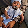 baby, infant, child, portrait, sitting, pacifier, baby_hat, blue_outfit, bib, onesie, smiling, wide_eyed, indoor, couch, adult, legs, family, close_up, soft_lighting, safe