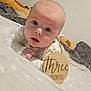 Martin is registered to the contest to win money with this photo: baby, infant, child, face, skin, blanket, wooden_sign, text, three_months, bed, soft_texture, clothing, eyes, mouth, head, pale_skin, indoors, portrait, cute, expression