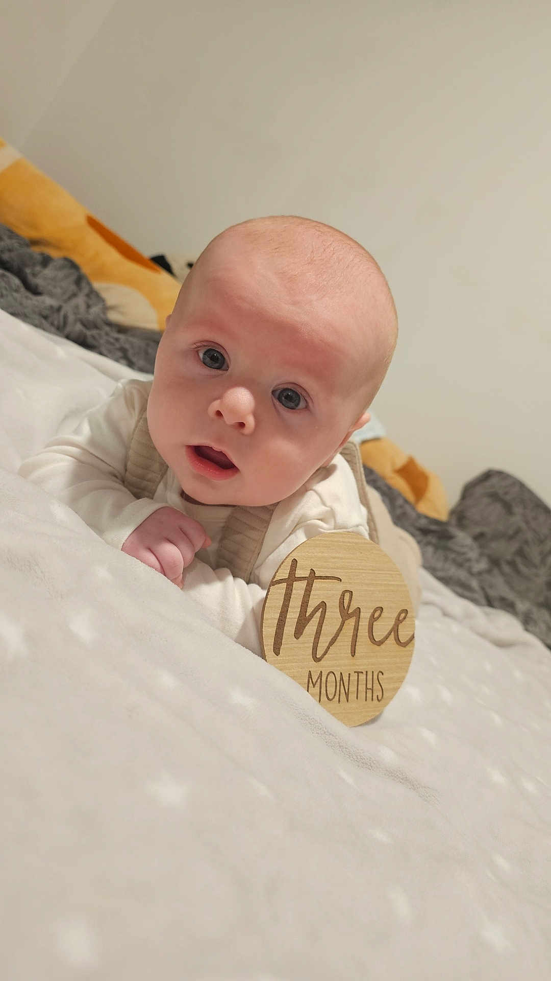 Martin is registered to the contest to win money with this photo: baby, infant, child, face, skin, blanket, wooden_sign, text, three_months, bed, soft_texture, clothing, eyes, mouth, head, pale_skin, indoors, portrait, cute, expression
