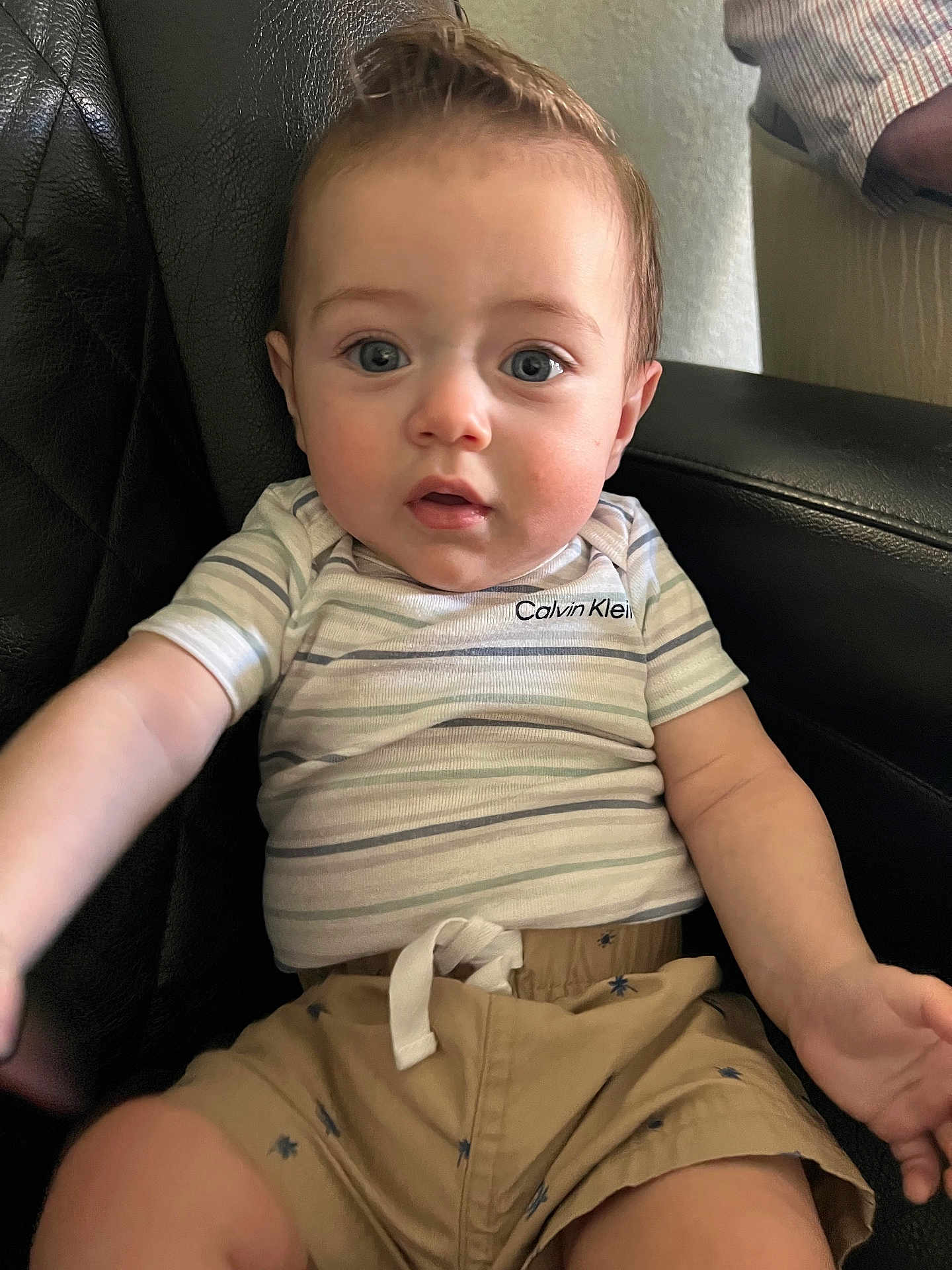 Kayden is registered to the contest to win money with this photo: baby, child, infant, calvin_klein, striped_shirt, beige_shorts, leather_chair, blue_eyes, short_hair, mohawk, cute, sitting, indoor, curious, person, apparel, casual_clothing, closeup, portrait, young