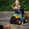 toddler, child, toy, dump_truck, tools, outdoor, mud, face_paint, barefoot, denim_overalls, play, curious, greenery, stone_patio, yellow, orange, black, plastic, expression, summer