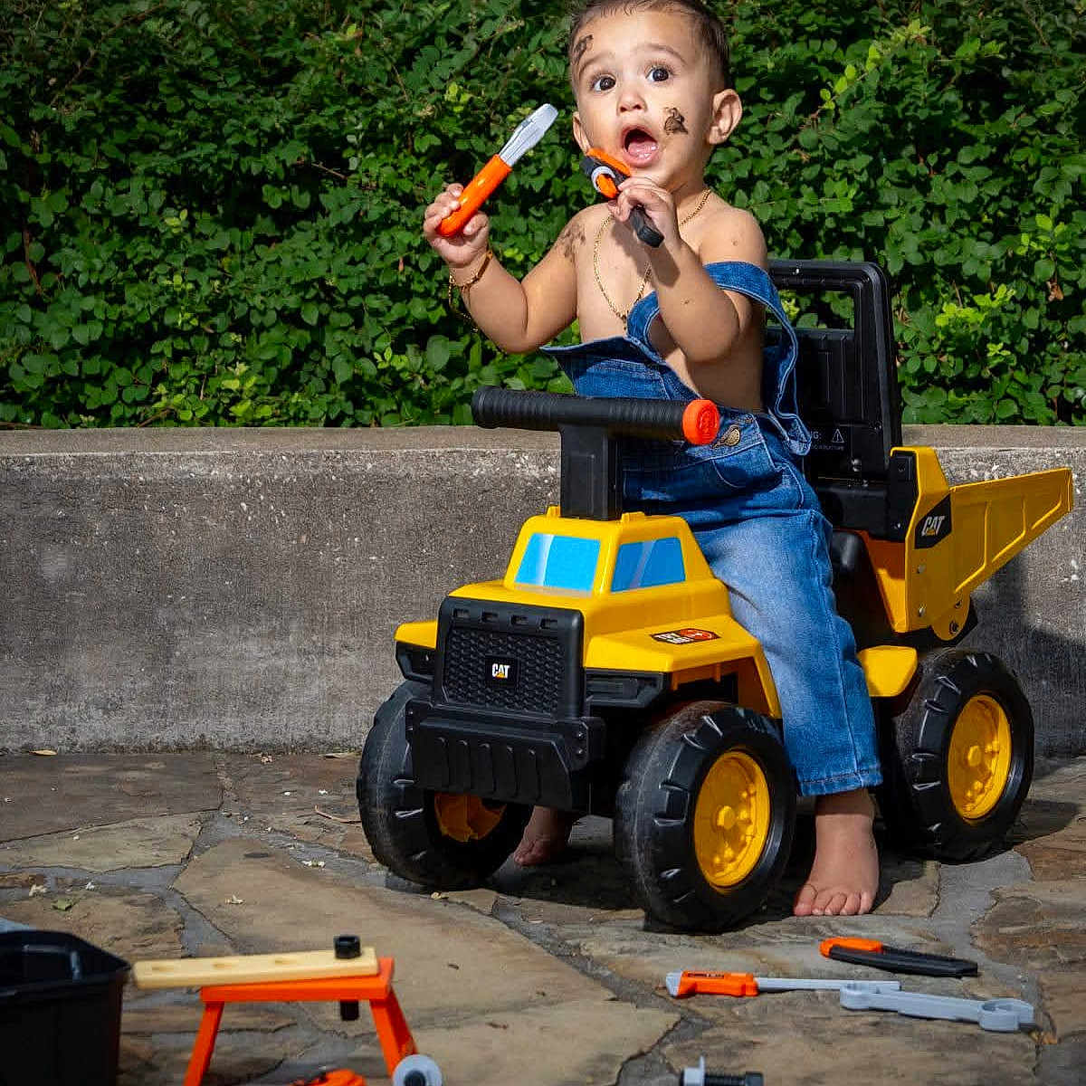 Neil joined the competition — help win amazing prizes! toddler, child, toy, dump_truck, tools, outdoor, mud, face_paint, barefoot, denim_overalls, play, curious, greenery, stone_patio, yellow, orange, black, plastic, expression, summer