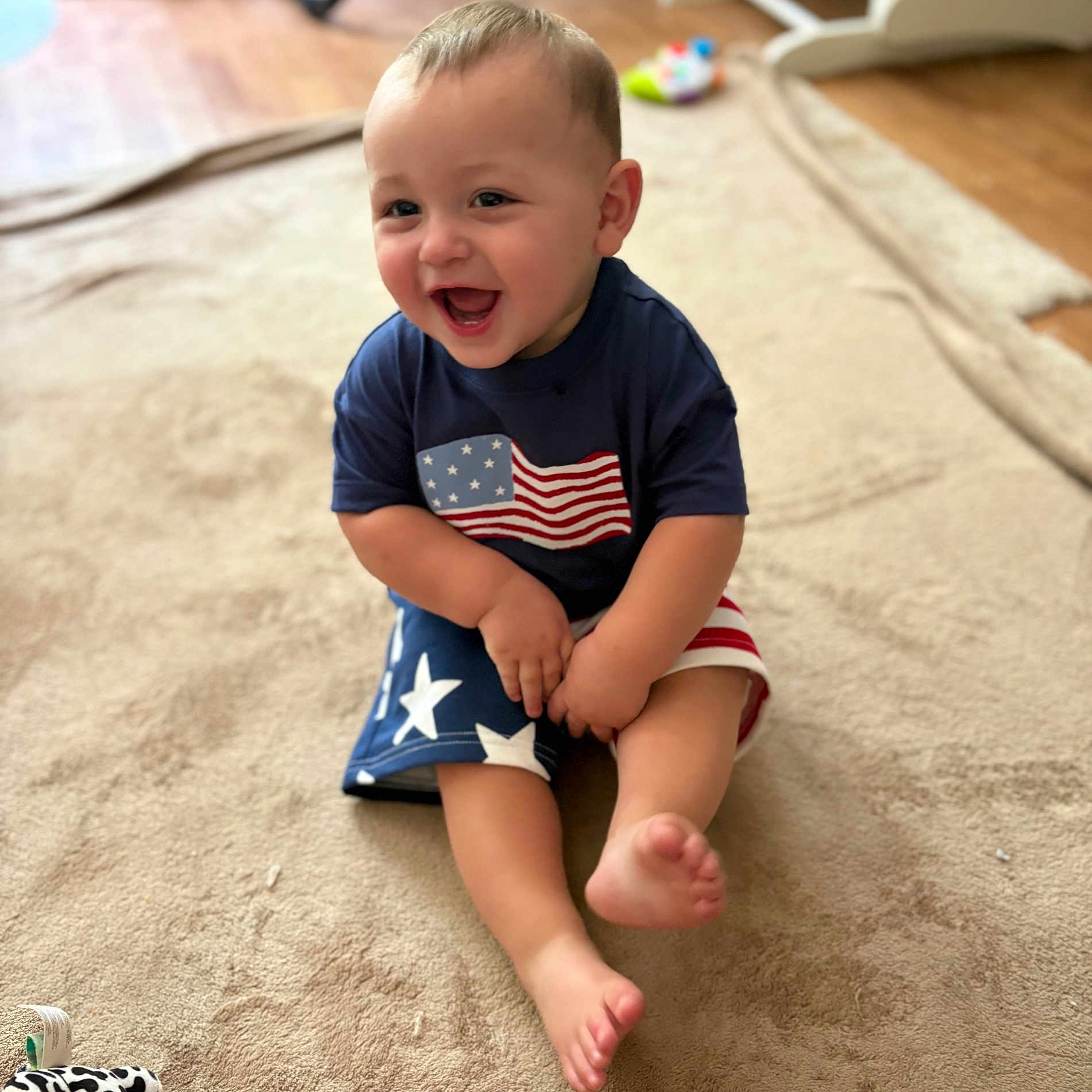 Aiden is registered to the contest to win money with this photo: toddler, child, smiling, happy, sitting, indoor, carpet, shorts, tshirt, american_flag, barefoot, baby, cute, playful, person, portrait, skin, face, legs, toes