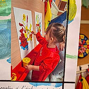 Lyvianah participe au concours pour gagner de l'argent avec cette photo : child, painting, red_smock, brush, artwork, paper, colorful_paint, yellow_paint_container, wooden_board, classroom, adult, creative_activity, indoor, young_child, hair_tied, hand, jeans, sneakers, playful, education