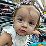 Sally is registered to the contest to win money with this photo: toddler, child, shopping_cart, toy, grocery_store, bow, hair_accessory, white_clothing, face, eyes, cute, portrait, indoor, person, small_child, shopping, retail, curious, young_child, casual