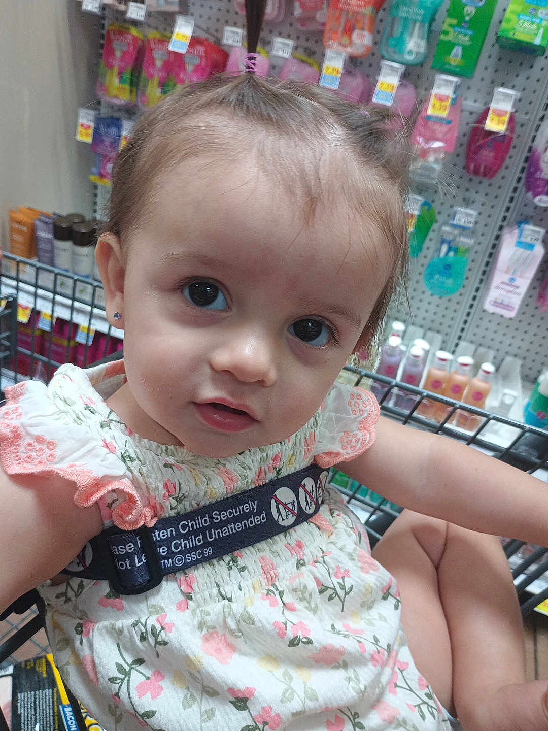 Sally is registered to the contest to win money with this photo: toddler, child, shopping_cart, floral_dress, safety_strap, store_shelves, products, curious_expression, earrings, hair_tie, indoor, retail, baby, person, cute, closeup, face, child_safety, grocery_store, shopping