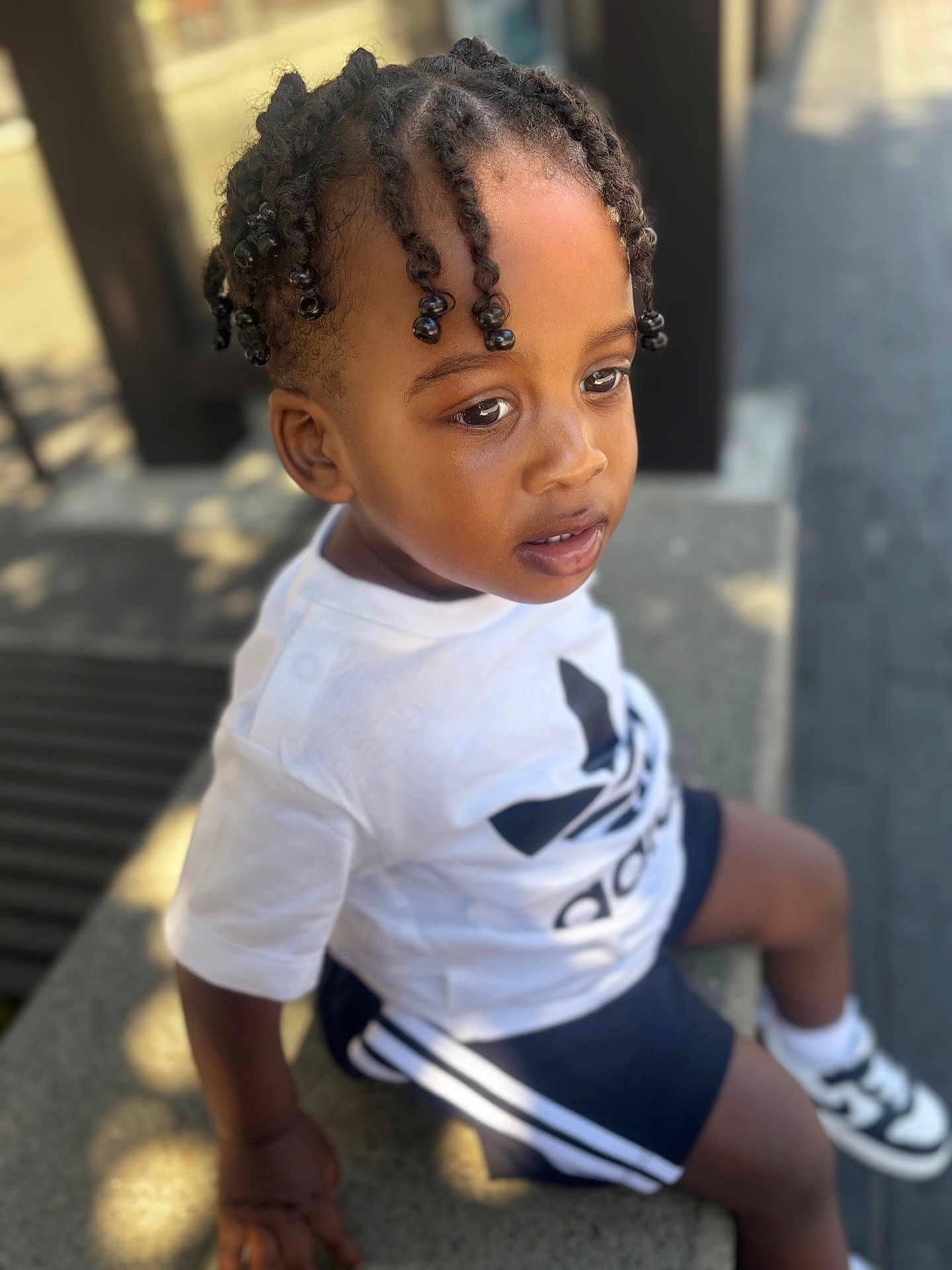 Kenzo participe au concours pour gagner de l'argent avec cette photo : toddler, child, braided_hair, white_tshirt, shorts, sneakers, outdoor, bench, sunlight, shadow, side_view, casual_clothing, portrait, young_child, curious, daylight, street, fashion, person, sitting