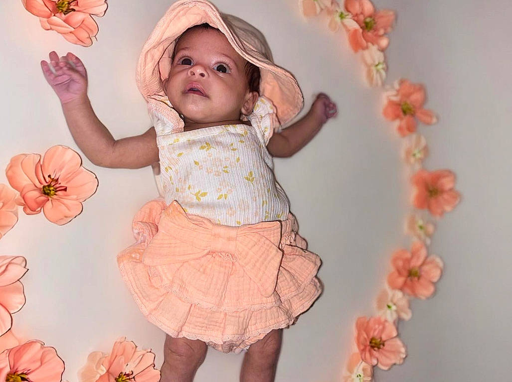 Tala is registered to the contest to win money with this photo: baby, baby_toddler_clothing, cheek, child, dress, flower, happy, headgear, headwear, pattern, peach, person, petal, pink, plant, product, ruffle, skin, sleeve, sun_hat