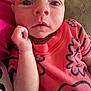 baby, infant, child, close_up, face, hand, orange_shirt, patterned_clothing, couch, indoor, living_room, bookshelf, dvd, curious, resting, wide_eyes, soft_furniture, portrait, young_child, casual_clothing