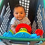 baby, child, shopping_basket, toy, steering_wheel, indoor, floor, curly_hair, clothing, play, bright_colors, plastic, cute, person, portrait, looking_up, expression, seat, hands, young