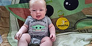 Leland is registered to the contest to win money with this photo: applique, baby, bodypart, clothing, couch, face, finger, furniture, hand, happy, head, homedecor, newborn, pattern, person, photography, portrait, shorts, sitting, t_shirt