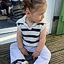 casual_clothing, child, curious_expression, curly_hair, cute, daylight, fashion, glass_door, outdoor, person, portrait, reflection, side_view, sitting, sneakers, striped_sweater, toddler, white_pants, wooden_deck, young_child