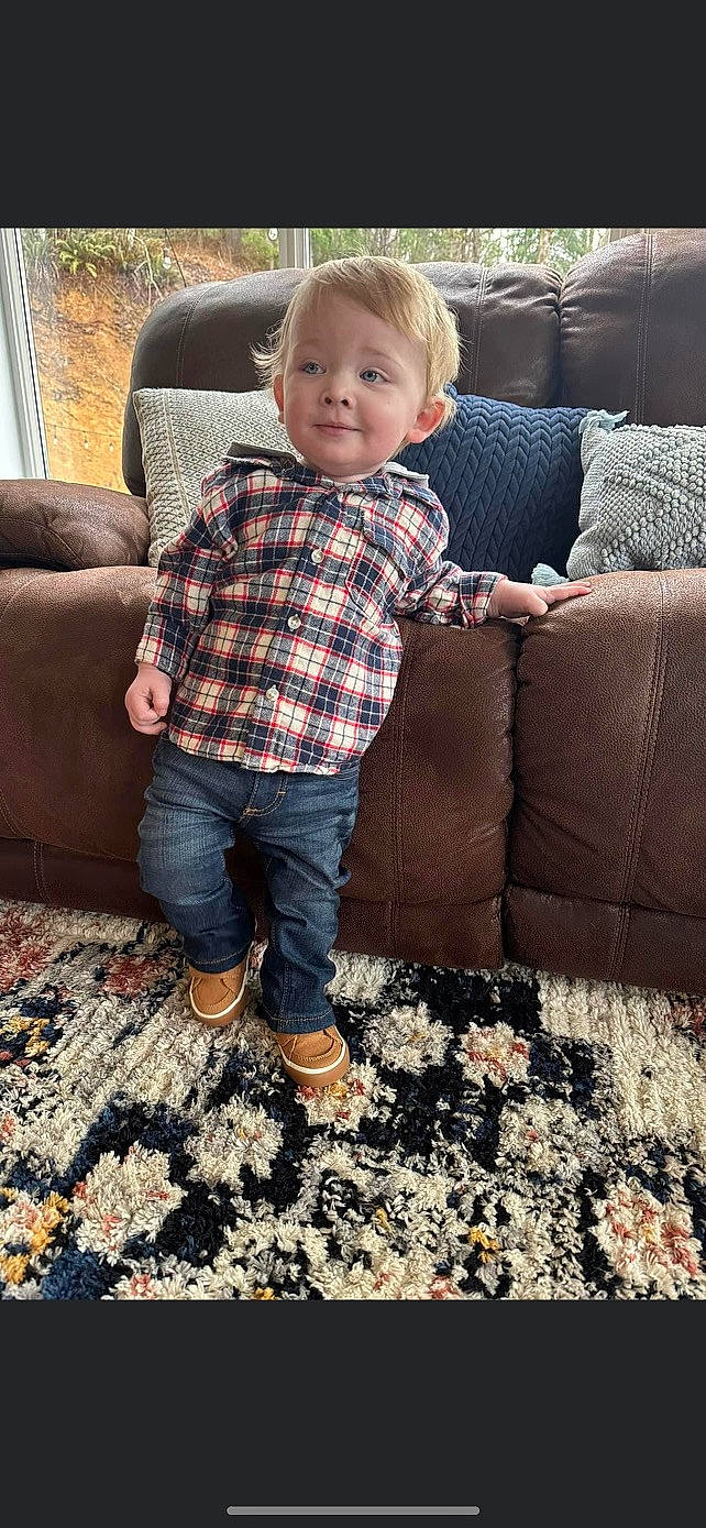 Graysen is registered to the contest to win money with this photo: baby, baby_toddler_clothing, child, comfort, couch, eye, flooring, fun, joy, living_room, pattern, person, plaid, room, sitting, sleeve, smile, studio_couch, tartan, textile