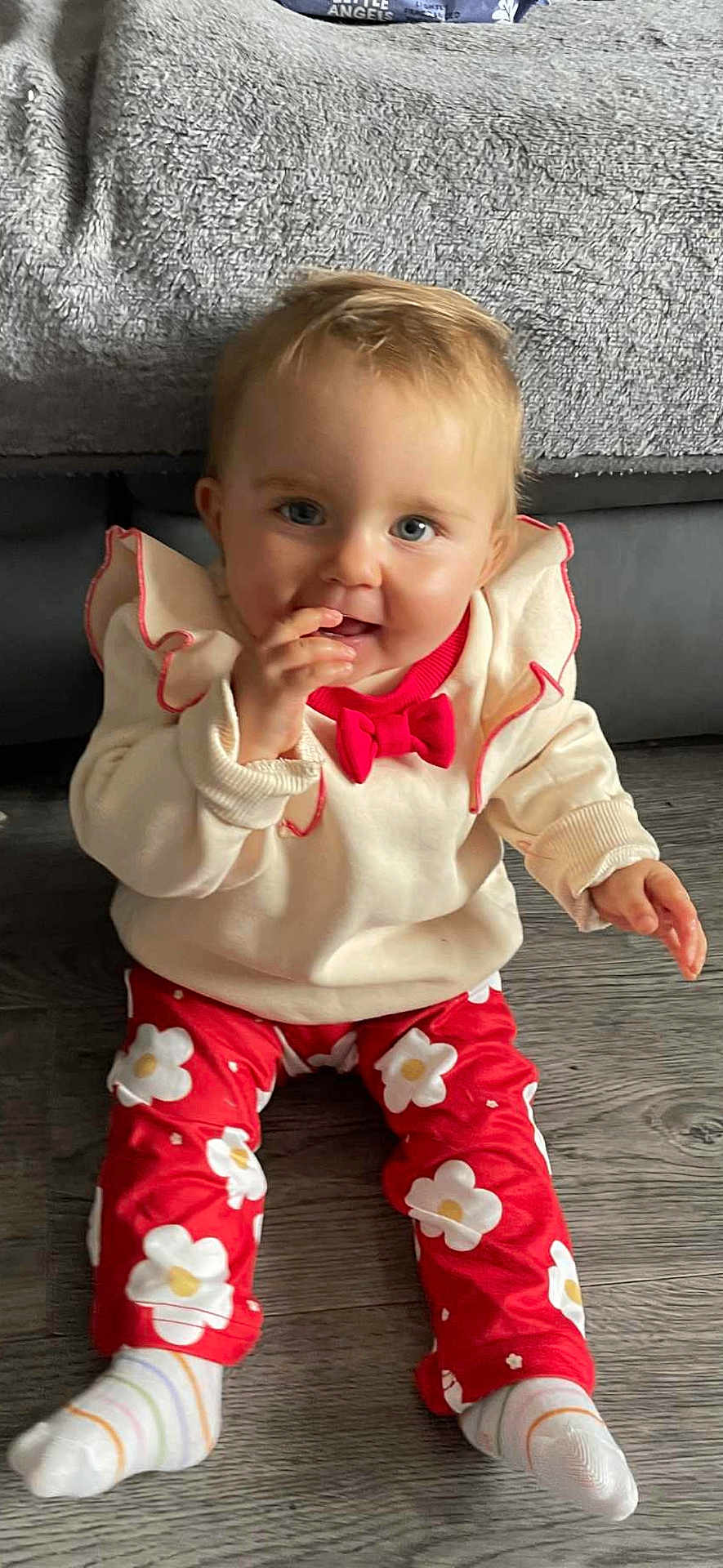 Brianna joined the competition — help win amazing prizes! baby, child, sitting, floor, red_pants, flower_pattern, cream_sweater, red_bow, sock, wooden_floor, indoor, person, cute, smiling, face, fingers, curious, cozy, blanket, couch