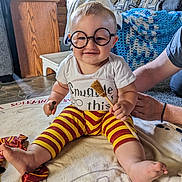 Anthony joined the competition — help win amazing prizes! baby, child, glasses, striped_pants, broomstick, blanket, smile, indoor, person, hand, floor, wooden_furniture, couch, decor, plaid_blanket, foot, toddler, cute, costume, happy